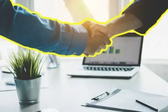 Two people shaking hands across a desk after agreeing business finance, with funding figures on a laptop.