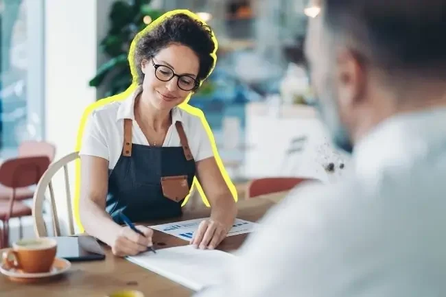 A business owner signs a commercial finance agreement while her broker talks her through the paperwork.
